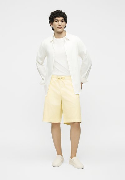 Άνδρας που στέκεται με τα χέρια στις τσέπες, φοράει λευκό πουκάμισο, λευκό μπλουζάκι, ανοιχτό κίτρινα σορτς με σκοινί και λευκά παπούτσια χωρίς κορδόνια.