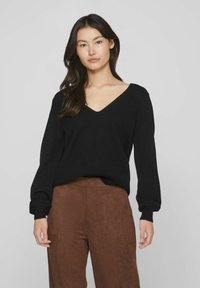 Suéter negro de pico con mangas largas y ligeramente abullonadas. Textura suave y corte relajado, combinado con pantalones marrones de ante.