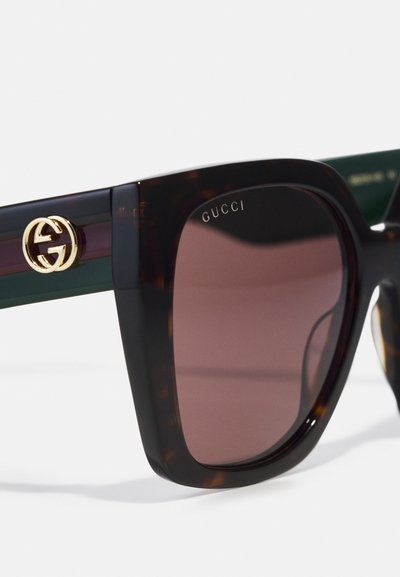 Gucci-zonnebril met een schildpadvormige montuur, donkerbruine glazen en een gouden logo-accent aan de zijkant. Heeft een rechthoekige vorm en groene poot.