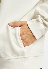 Fehér kapucnis pulóver elülső zsebből és bordás mandzsettákból. Lágy szövettextúra látható. Kéz a zsebben a méretérték kedvéért.