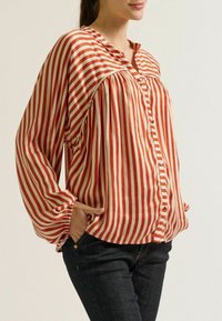 Rood en crème gestreepte blouse met een losse pasvorm, geplooide details, knoopsluiting en lange, elastische mouwen. Gecombineerd met donkere jeans.