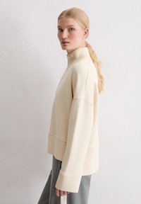 Beżowa bluza z wysokim kołnierzem o prążkowanej fakturze, oversize'owy krój i skrócony krój dołu, w połączeniu z szarymi spodniami. Gładki materiał, minimalne wykończenie.