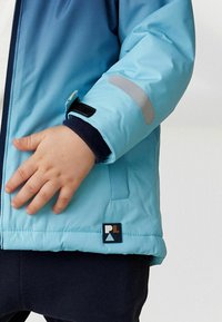 Veste isolante bleu clair avec des accents navy, dotée d'une fermeture éclair, de poignets élastiques et d'un patch logo. Texture lisse, conçue pour la chaleur.