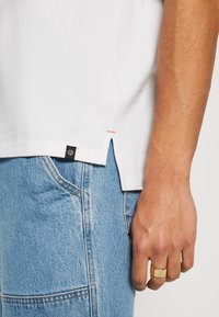 Tricou alb din bumbac, cu slituri laterale, având o cusătură de accent portocalie și o mică etichetă cu logo negru. Asortat cu blugi din denim albaștri.