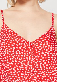 Vestido rojo de lunares blancos, con tirantes ajustables, escote en V y un diseño de botones en la parte frontal. Textura suave.