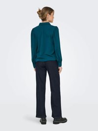 Blusa lunga teal con colletto, spalle a palloncino e polsini abbottonati, abbinata a pantaloni neri a gamba larga e scarpe nere.