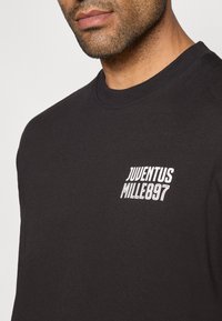 T-shirt de algodão preto com gola redonda, apresentando texto bordado em branco "JUVENTUS MILLE897" no lado superior esquerdo. Textura suave, corte standard.