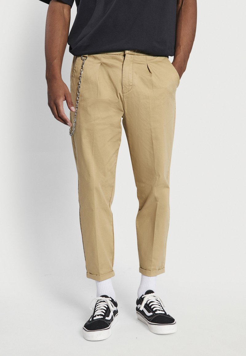 Beige chinos with a relaxed fit, featuring side pockets, a zipper fly, and a silver chain detail. Paired with black and white sneakers.