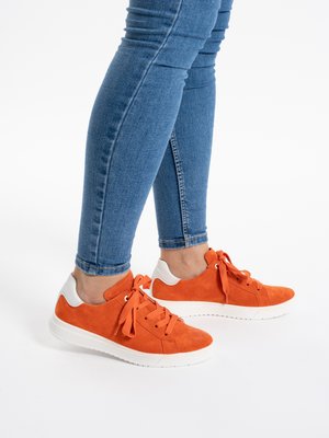 Person wearing blue skinny jeans and bright orange sneakers with white soles, standing on a white floor.