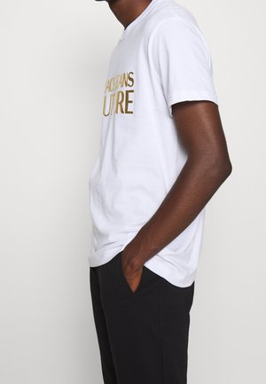 Biała bawełniana koszulka z złotym haftowanym napisem "FUTURE", z okrągłym dekoltem i krótkimi rękawami, w zestawieniu z ciemnymi spodniami.