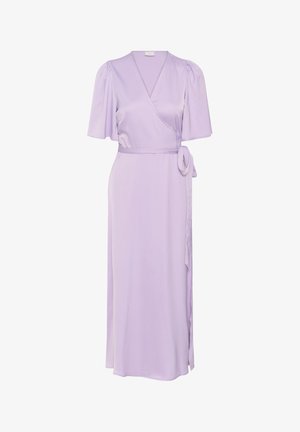 Femme portant une robe portefeuille violet clair avec des manches mi-longues et des talons noirs ouverts, debout devant un fond clair uni.