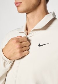 Nike zip-up jacket made of lightweight beige fabric, featuring a black swoosh logo, stand collar, and elastic cuffs; hand gripping the zipper.