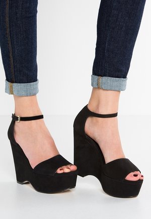 Pieds portant des talons compensés en daim noir avec des brides à la cheville et un jean skinny bleu foncé retroussé sur un fond blanc.