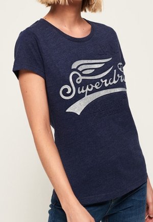 Kvinde iført en mørkeblå Superdry t-shirt med hvidt vinget logo og tekst, kombineret med mørkeblå jeans.