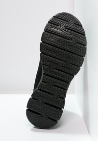 Černá podrážka bot Skechers s hlubokými drážkami a textura pro lepší uchopení, nakloněná proti bílému rohu pozadí.