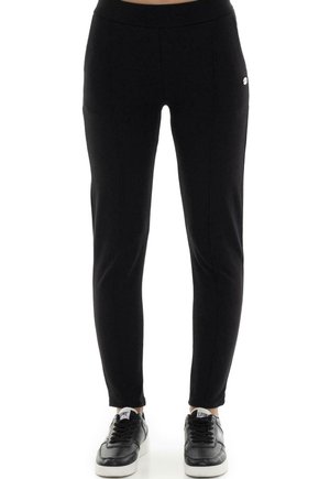 Pantalon noir fuselé avec une large ceinture et un petit logo, porté avec des baskets noires sur fond blanc.