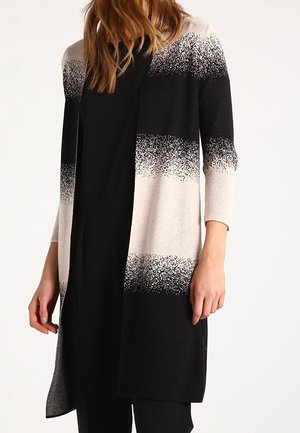 Kobieta ubrana w długi kardigan w czarno-beżowym gradiencie, na czarnym topie i spodniach, stojąca na jasnym, jednolitym tle.