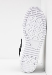 Schwarzer Wildleder-Sneaker mit einer weißen Gummisohle, die ein geometrisches Profilmuster aufweist. Enthält ein Logo auf der Sohle und metallene Ösen für die Schnürsenkel.