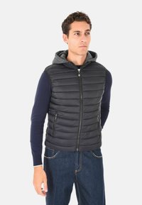 Uomo che indossa un gilet trapuntato nero con cappuccio grigio sopra una maglietta a maniche lunghe blu navy e jeans blu, in piedi davanti a uno sfondo chiaro e semplice.