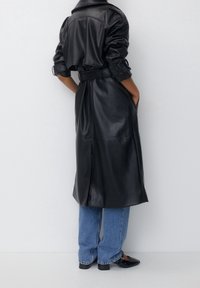 Personne portant un trench-coat en cuir noir ceinturé, sur un jean bleu et des chaussures noires à bout pointu, se tenant devant un mur blanc uni.