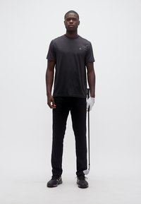 Άνδρας ντυμένος με μαύρη γκολφ ενδυμασία και λευκό γάντι, κρατώντας ένα γκολφ ρόπαλο, στέκεται μπροστά σε λευκό φόντο.