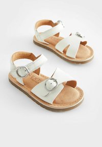 Vita läder sandaler med en brun mockainsocka, med två justerbara remmar och en rund silverfärgad spänne. Mjuk, platt sula.