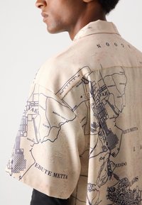 Kortsleeved skjorte laget av glatt stoff med en beige bakgrunn og et detaljert trykket kart i mørkeblått, som viser ulike steder.