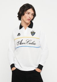 Camisa polo blanca de manga larga con cuello y puños negros. Presenta rayas amarillas y azules en el pecho y un logotipo y texto bordados en negro.