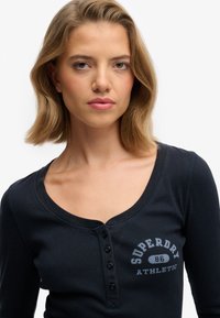 Haut henley à manches longues bleu marine en coton côtelé, avec un col arrondi et une patte de boutonnage ornée du graphisme "Superdry Athletic".