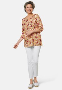 Florale Tunika in Gelb, Rot und Lila mit V-Ausschnitt und Dreiviertelärmeln, kombiniert mit weißen Hosen und hellen Espadrilles.