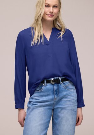 Donna bionda che indossa una camicetta a maniche lunghe blu reale infilata in jeans azzurri con una cintura nera, in piedi davanti a uno sfondo semplice.