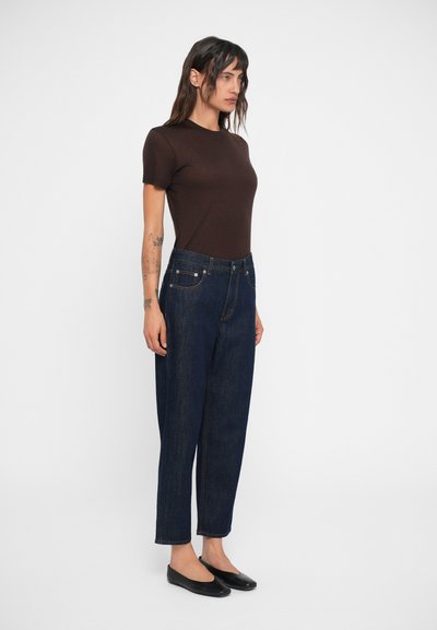 Camisa de manga corta marrón oscuro combinada con jeans de mezclilla oscuros de cintura alta. Los jeans tienen un diseño clásico de cinco bolsillos y costuras claras. Bailarinas negras.