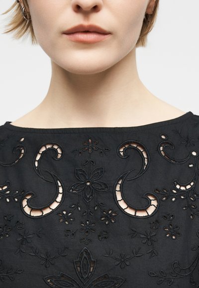 Zbliżenie na dolną część twarzy i szyję osoby ubranej w czarną bluzkę z haftem w formie kwiatowych wzorów i zawijasów z wycięciami.