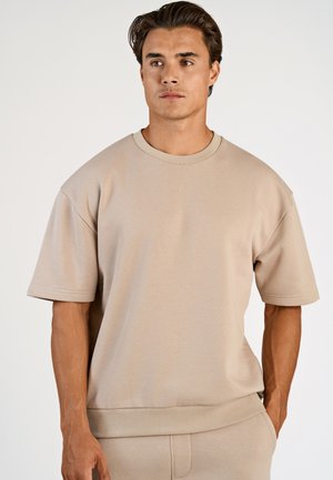 Mann trägt beige kurzärmliges Sweatshirt und passende Hose, steht mit Händen in den Taschen und schaut leicht nach rechts vor einem schlichten Hintergrund.