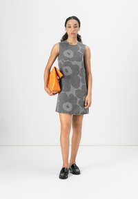 Sivá šatka bez rukávov s veľkým kvetinovým vzorom, z denimu, so strapcovým lemom. Modelka drží oranžovú kabelku a nosí čierne mokasíny.