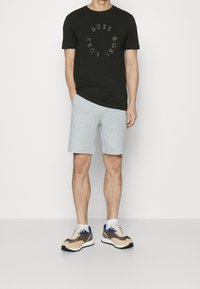 Man som bär svart T-shirt med cirkulär "BOSS" text, ljusgrå shorts, vita strumpor och beige och bruna sneakers, står med ena handen i fickan.