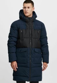 Abrigo acolchado azul marino con un panel negro en el pecho, dos bolsillos frontales, cierre con cremallera y capucha desmontable. Presenta una textura acolchada.