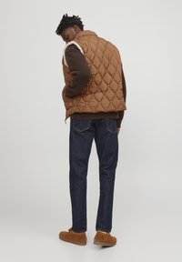 Quilted brown puffer vest, fleece-lined shoulders, paired with dark denim jeans and brown suede shoes, viewed from the back.