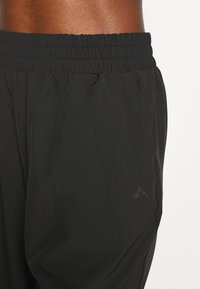 Mustat urheiluhousut, joissa on joustava vyötärönauha. Ne on valmistettu sileästä, kevyestä kankaasta ja sivussa on hillitty logodetalji.