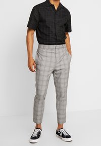 Chemise noire à manches courtes avec motif à carreaux et accents marron, associée à un pantalon fuselé à carreaux gris clair et des baskets noires.