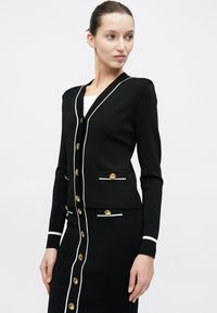 Μαύρο ριμπ πλεκτό ζακέτα με λευκή διακοσμητική ραφή, χρυσά κουμπιά και δύο μπροστινές τσέπες. Συνδυάζεται με μαύρη φούστα σετ με παρόμοιες λεπτομέρειες.