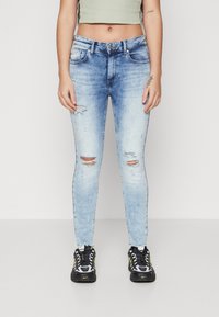 Jeans skinny desgastados de color azul claro con áreas descoloridas, desgastes en las rodillas y un diseño estándar de cinco bolsillos. Acompañados de zapatillas deportivas negras y amarillas.