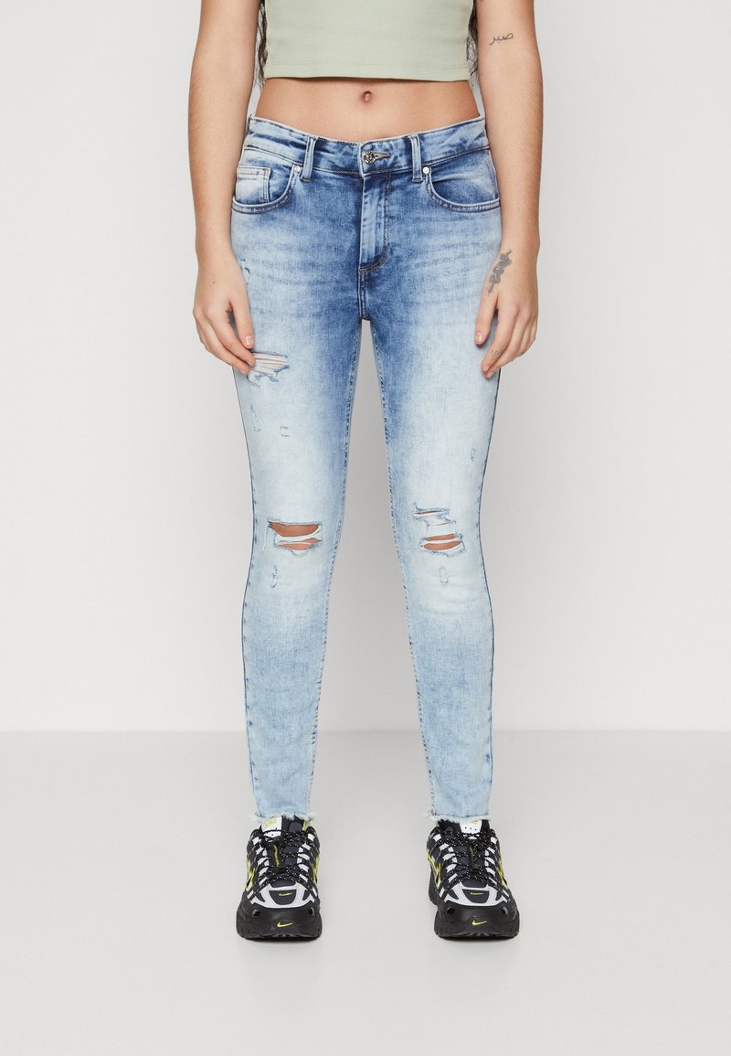 Jeans skinny desgastados de color azul claro con áreas descoloridas, desgastes en las rodillas y un diseño estándar de cinco bolsillos. Acompañados de zapatillas deportivas negras y amarillas.