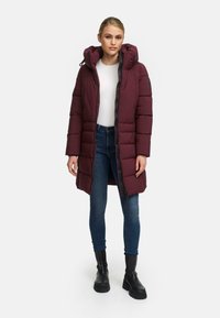 Bordeaux pufferjas met een afneembare capuchon, ritssluiting en een horizontaal gestikt patroon. Draagt over een witte top met blauwe jeans.
