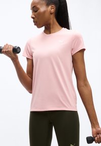 Γυναίκα με ροζ πουκάμισο και σκούρο κολάν που σηκώνει βαράκια 1 κιλού, γυμνάζεται μπροστά σε απλό λευκό φόντο.