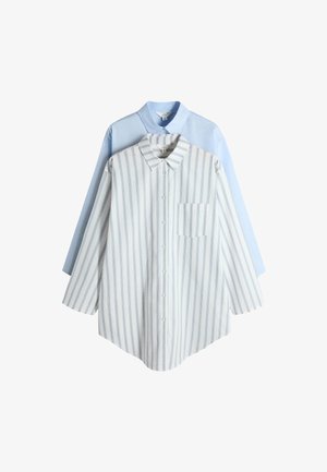 Vrouw staand met handen op de heupen, draagt een lichtblauw oversized overhemd en bruine sokken tegen een effen witte achtergrond.