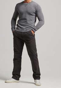 Mann in einem enganliegenden grauen Pullover, schwarzen Hosen und weißen Sneakers, der mit den Händen in den Taschen vor einem schlichten Hintergrund steht.