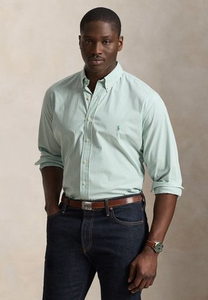 Άνδρας με πράσινο και λευκό ριγέ πουκάμισο με κουμπιά και γυρισμένα μανίκια, σκούρο τζιν, καφέ ζώνη και καφέ ρολόι με πράσινο καντράν.