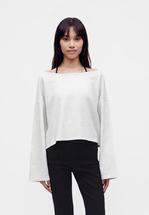 Jeune femme aux longs cheveux foncés portant un haut blanc ample à épaules dénudées avec manches longues et un pantalon noir, debout devant un fond clair uni.