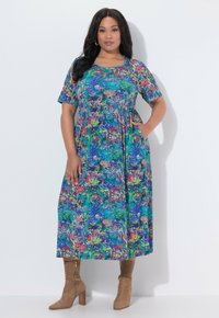 Kvinde står op ad en hvid væg iført blå blomsterprint midi-kjole med korte ærmer og tanfarvede ruskindsstøvler med hæl, den ene hånd i lommen.